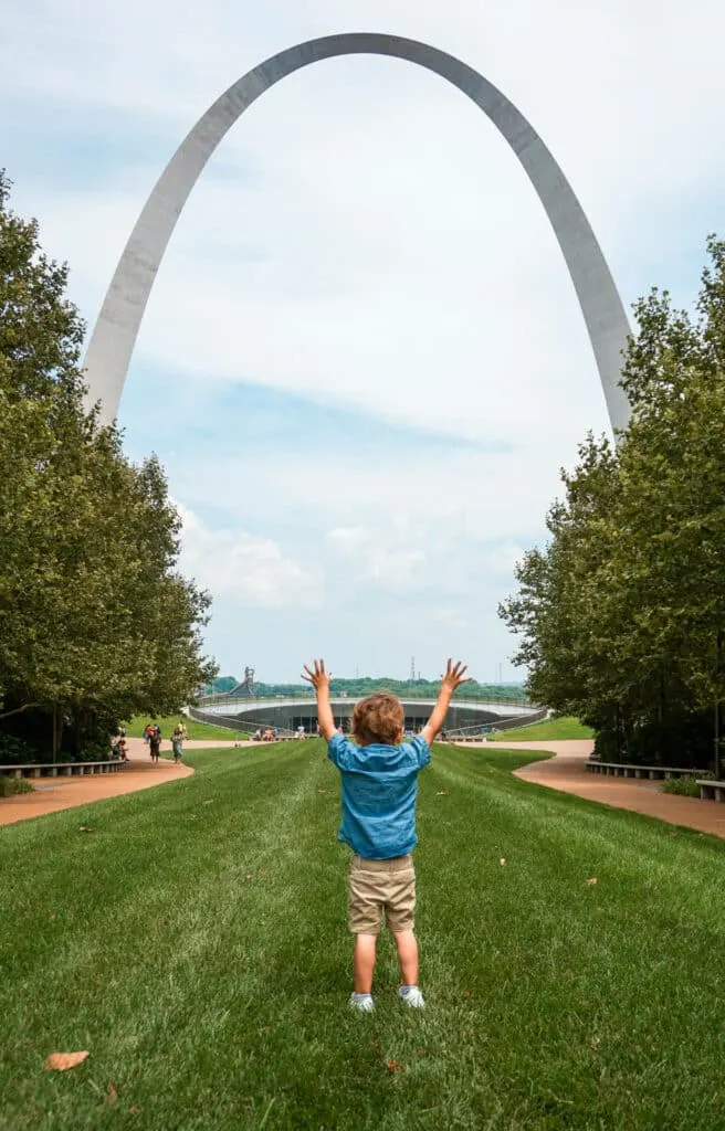 A boy facing the Gateway Arch while raising his arms.