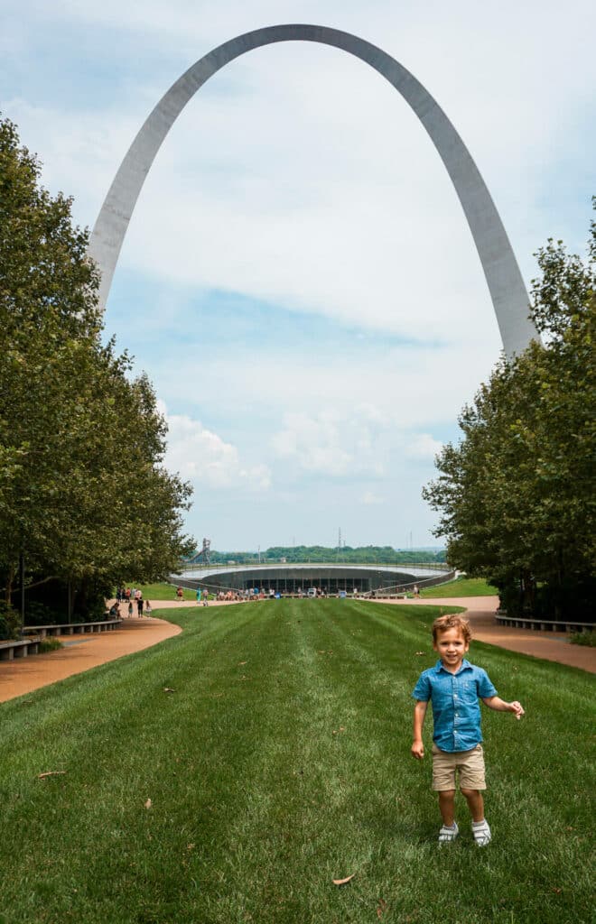A toddler boy on a green lawn with the Gateway Arch towering behind him.