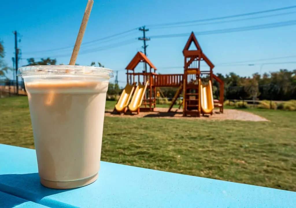 A cold latte drink with a wooden playground in the background located at FairLane Coffee and Cocktails in Dripping Springs. 