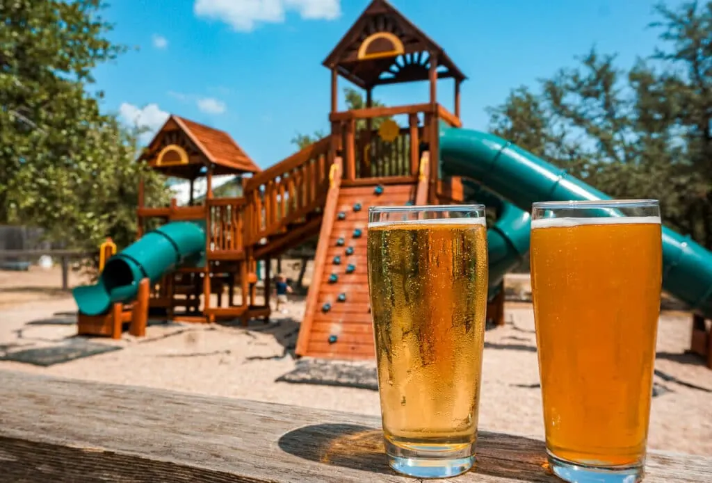 Two glasses of beer with a wooden playground in the background located at Family Business Beer Co. - a kid-friendly brewery in Drippings Springs