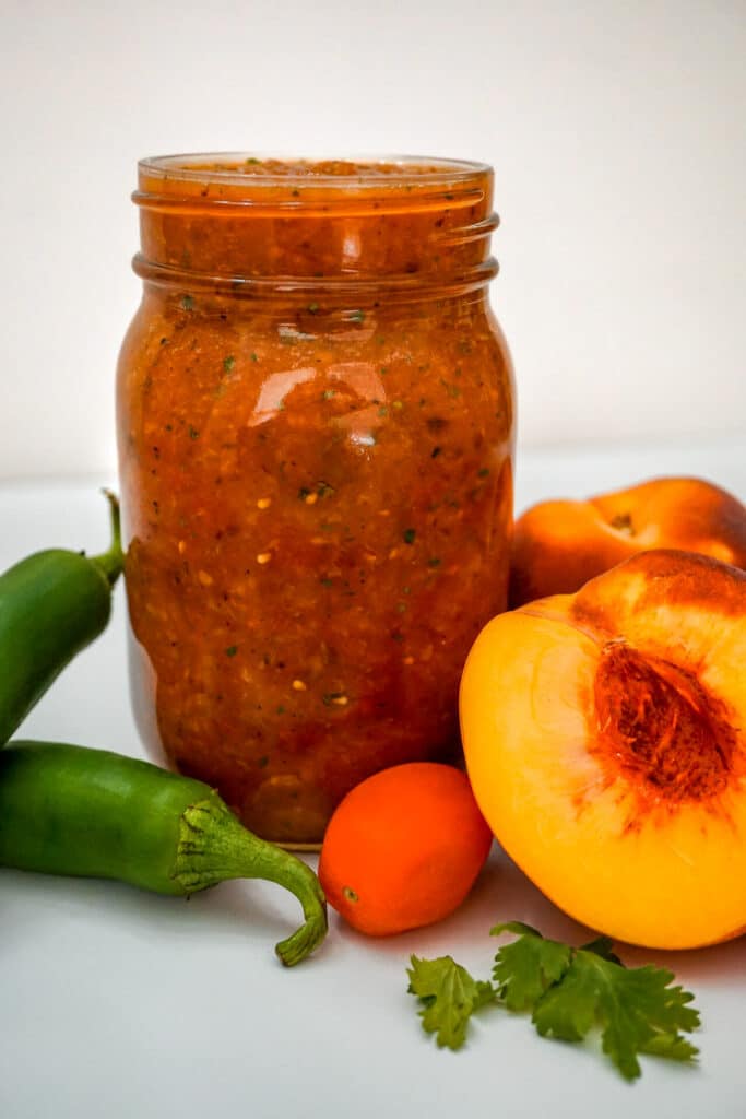 A major jar of Grilled Peach Salsa with jalapeños, cherry tomatoes, cilantro, and half a peach beside the jar.