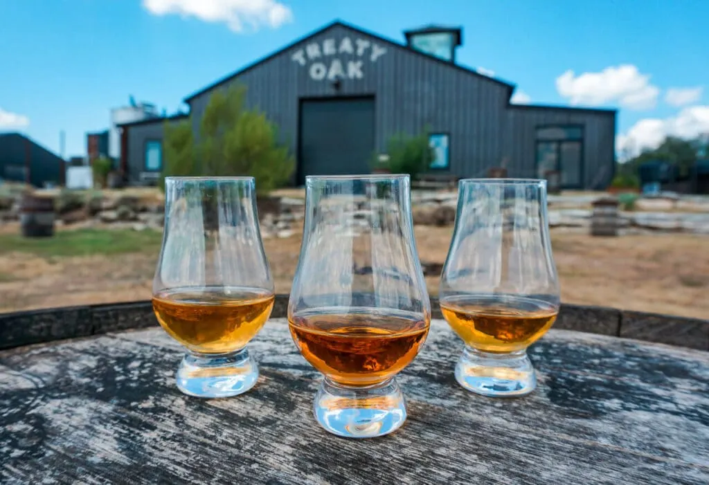 Three small glasses of a bourbon tasting with a massive barn shed in the background from Treaty Oak Distillery.