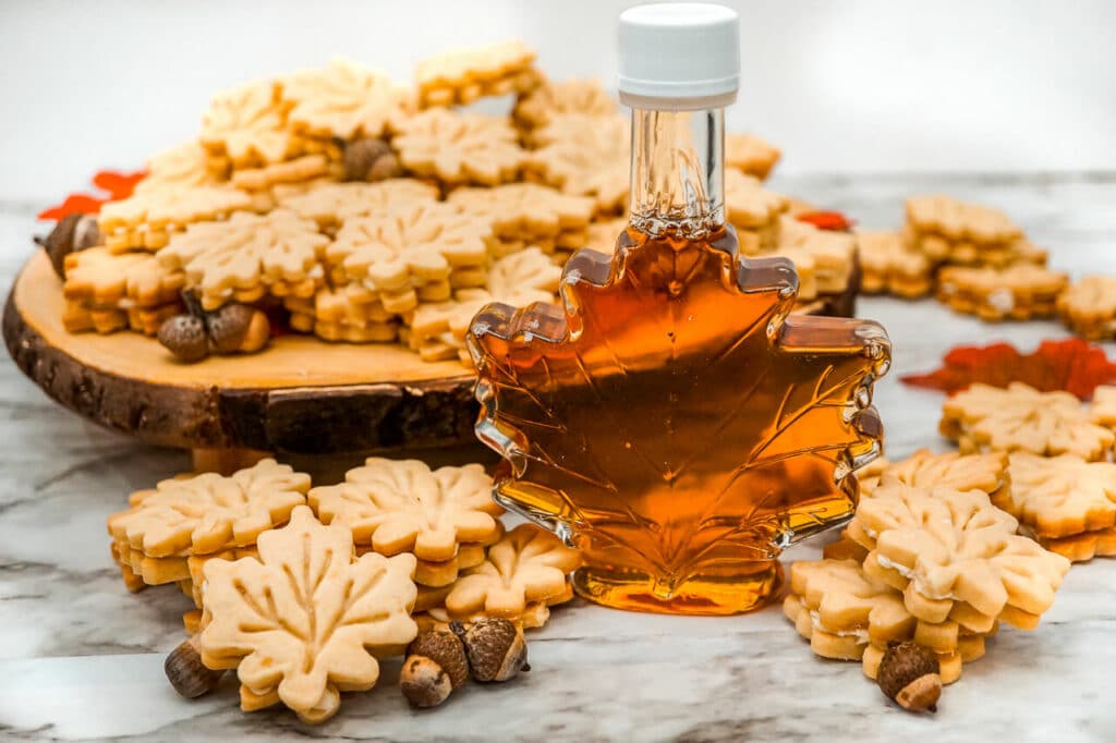 A bottle of pure maple syrup surrounded by Maple Leaf Cookies and a stack of the cookies on a plate behind the bottle.