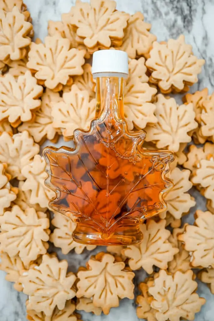 A bottle of pure maple syrup shaped like a maple leaf on top of Maple Leaf Cream Cookies.