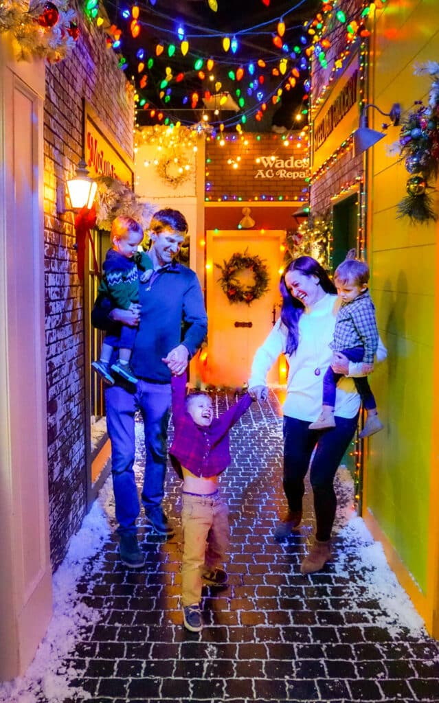 A family with three young boys smiling and having fun under a string of colorful Christmas lights at SNOWDAY - one of the best Christmas things to do in Dallas.