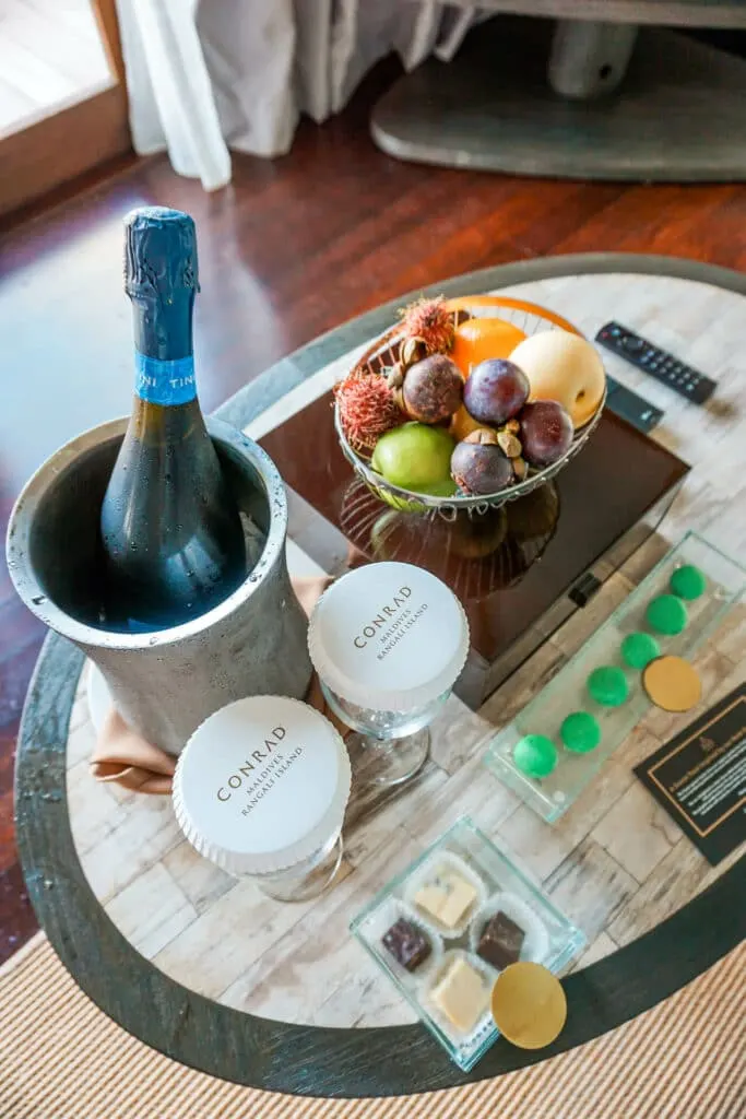 A table of amenities as a Hilton Honors Diamond Member from Conrad Maldives Rangali Island. A bottle of sparkling wine, chocolate truffles, green macarons, and a bowl of fresh fruit.