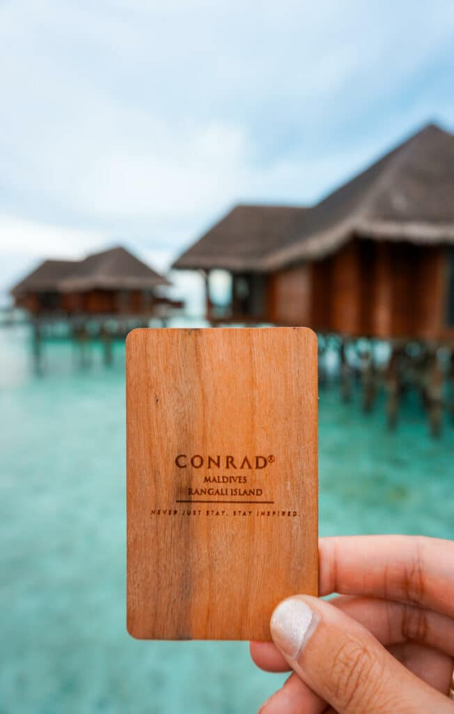 A brown key card with overwater villas in the background from my Conrad Maldives Rangali Island review.