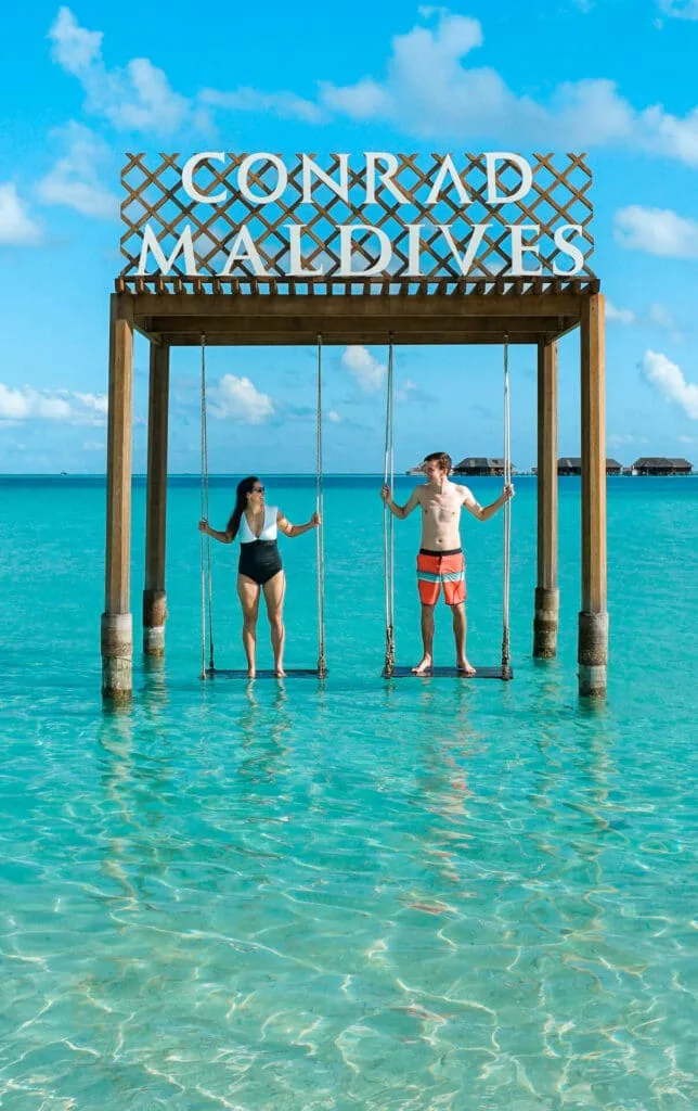 A couple standing on wooden swings in the middle of the ocean at Conrad Maldives Rangali Island.