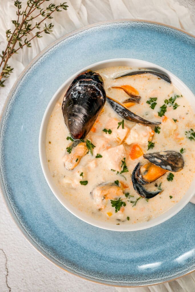 A bowl of Irish Seafood Chowder with mussels, chunks of cod and salmon, and chopped parsley on top of a light blue plate.