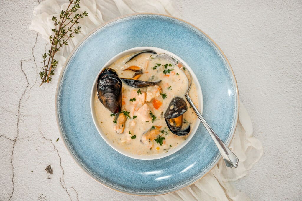 A sprig of fresh thyme next to a light blue plate with a bowl of Irish Seafood Chowder with open mussels, chunks of cod, and salmon. 