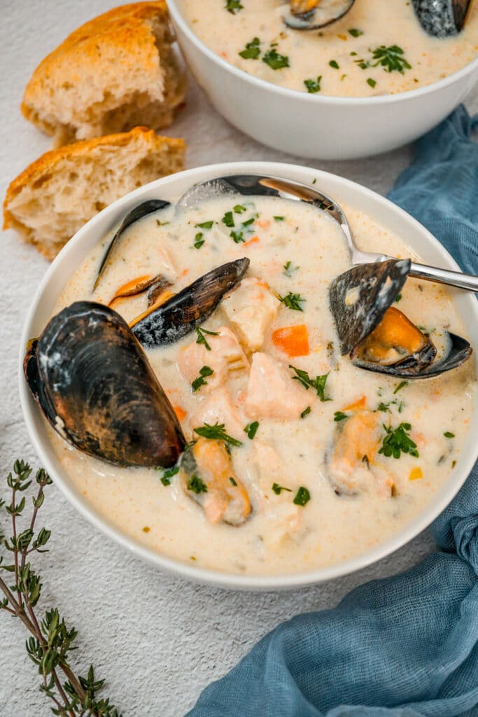 A spoon going into a bowl of Irish Seafood Chowder with mussels, chunks of cod and salmon, and chopped parsley on top. A blue cheesecloth napkin, sprig of thyme, and chunks of bread are surrounding the bowl of chowder. 