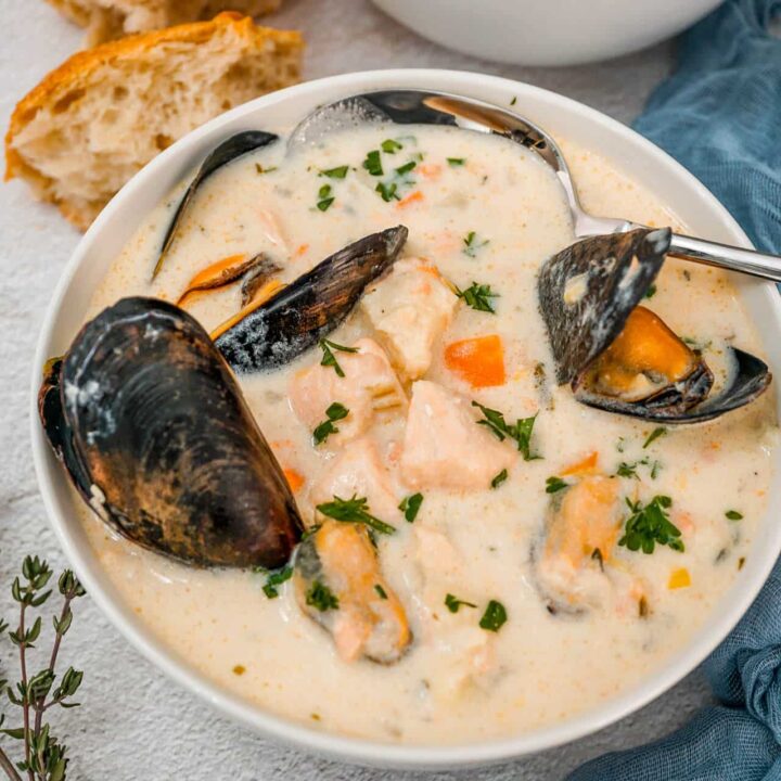 A spoon going into a bowl of Irish Seafood Chowder with mussels, chunks of cod and salmon, and chopped parsley on top. A blue cheesecloth napkin, sprig of thyme, and chunks of bread are surrounding the bowl of chowder.