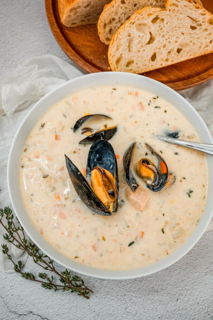 A spoon going into a bowl of Irish Seafood Chowder with open mussels, chunks of cod, and salmon. A plate of sliced bread and a fresh sprig of thyme next to the bowl.