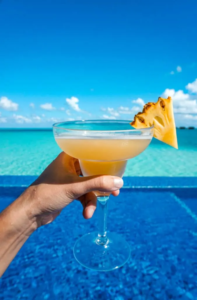 A hand holding a yellow cocktail with a slice of pineapple at the Quiet Pool at Conrad Maldives Rangali Island.