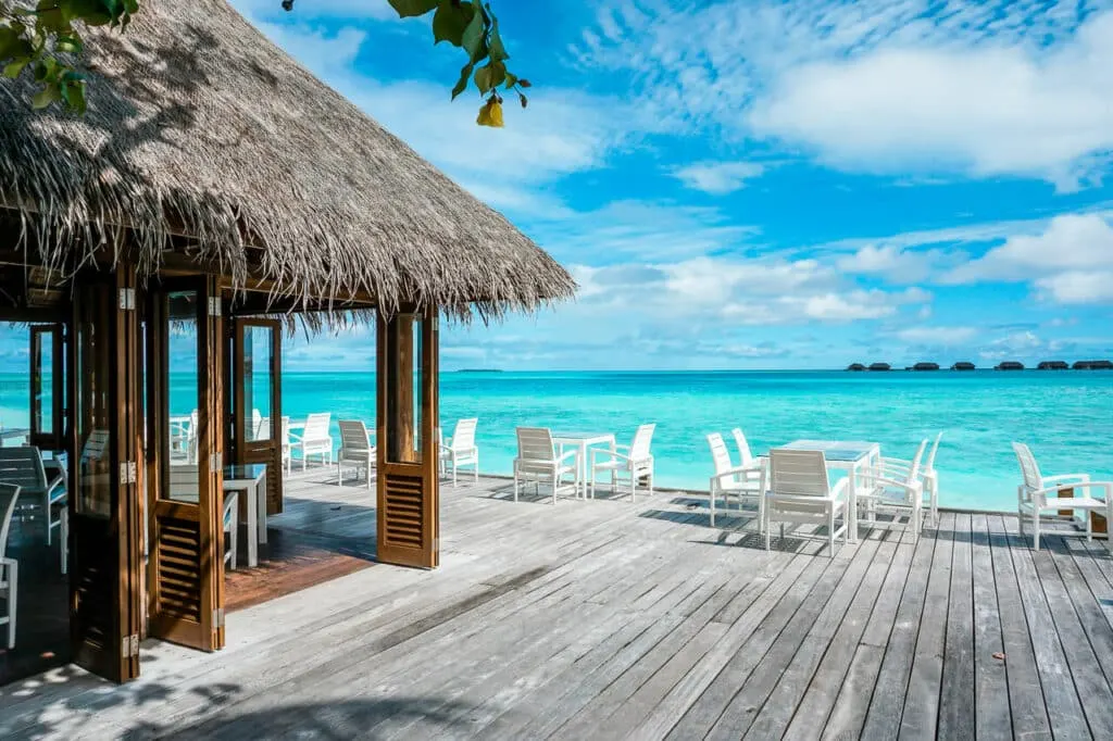 A large deck with a white dining tables and chairs and a thatch-roof restaurant overlooking a lagoon at the Conrad Maldives Rangali Island.