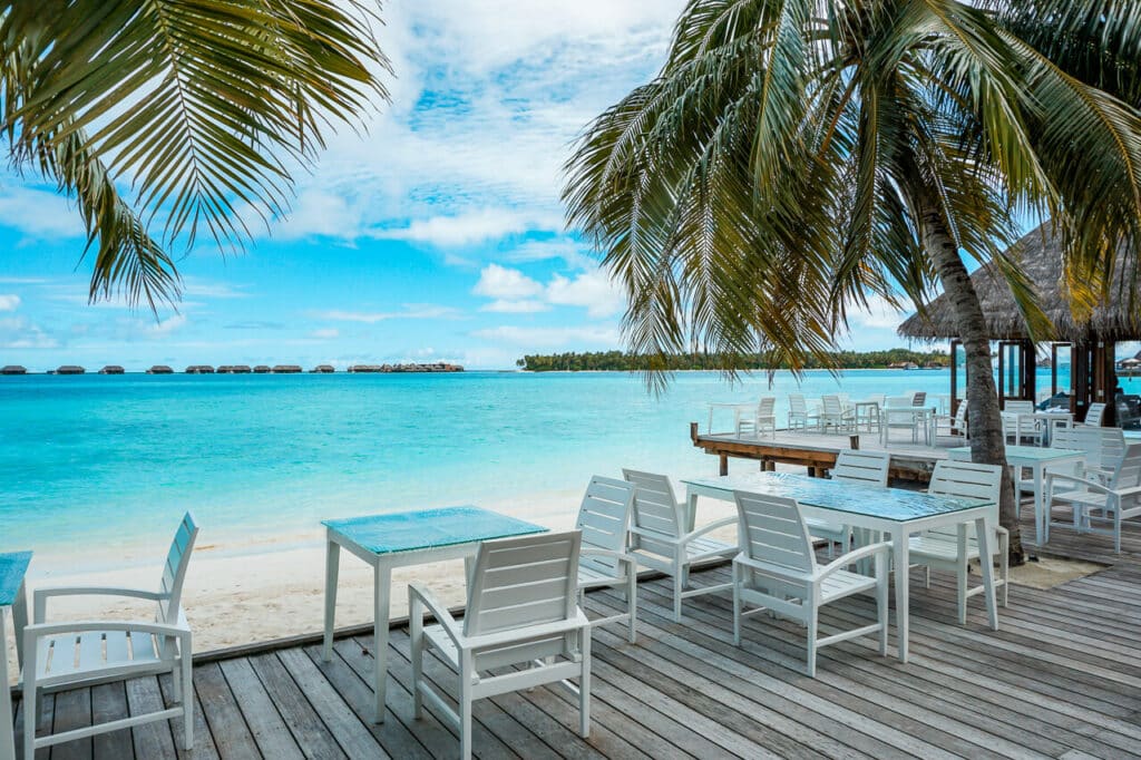 A few tables on a wooden deck with palm trees and the view of the Indian Ocean and overwater villas in the distance. Vilu Restaurant at Conrad Maldives Rangali Island. 