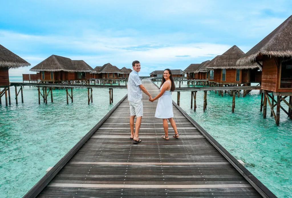 A couple dressed in white holding hands on a boardwalk with overwater villas at the Conrad Maldives Rangali Island.