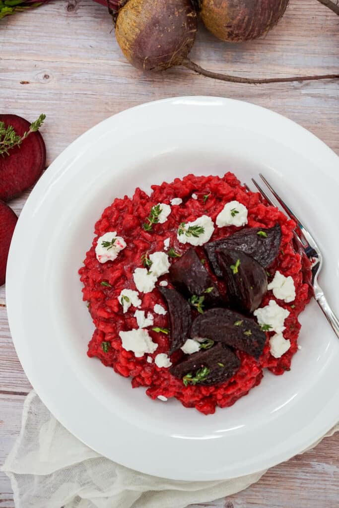A fork going into a white bowl of Roasted Beet Risotto with Goat Cheese crumbled on top and fresh thyme. 