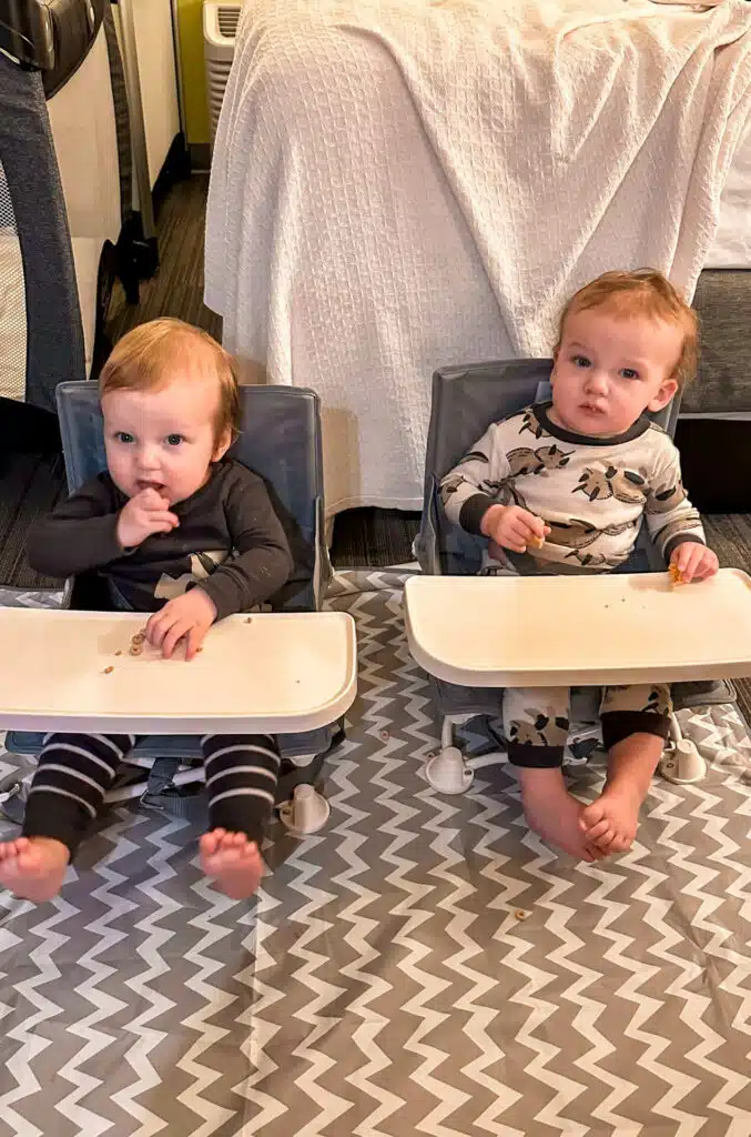 Twin boys sitting in travel high chairs in a hotel room waiting for their food.
