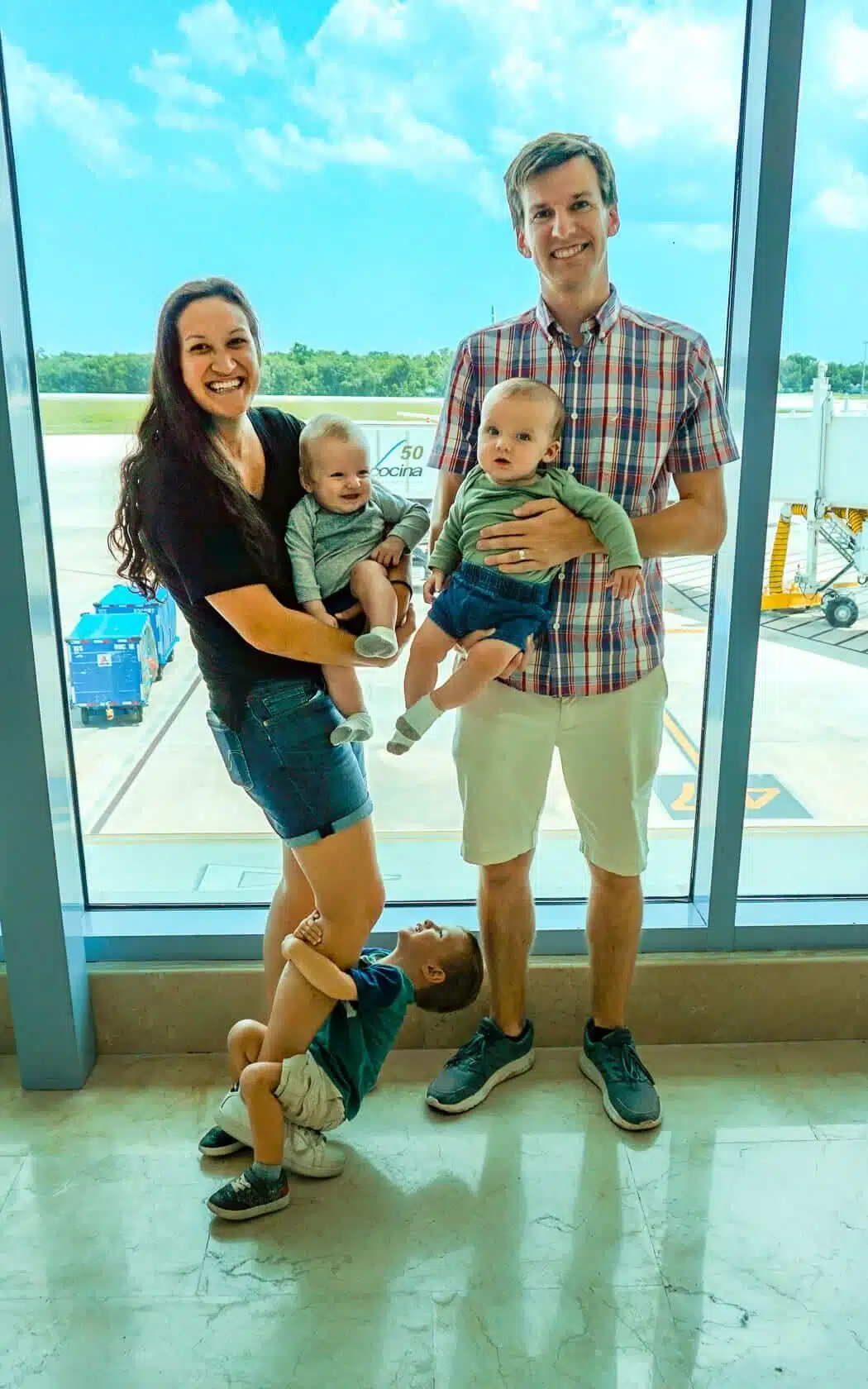Parents traveling with twins and a toddler hanging on his mom's legs at the airport.