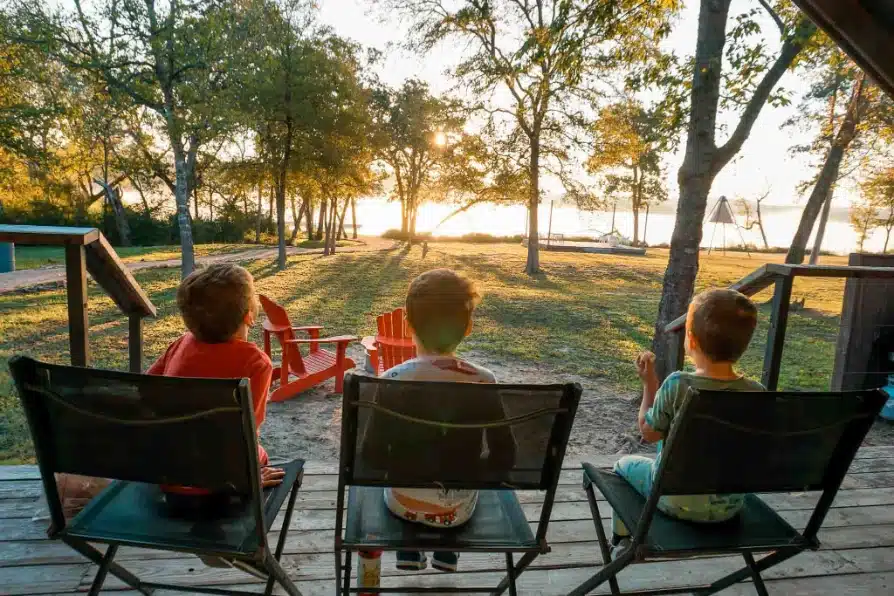 Three boys sitting on chairs on a patio looking out towards the sun shining on Lake Bastrop at Lake Bastrop North Shore Park.