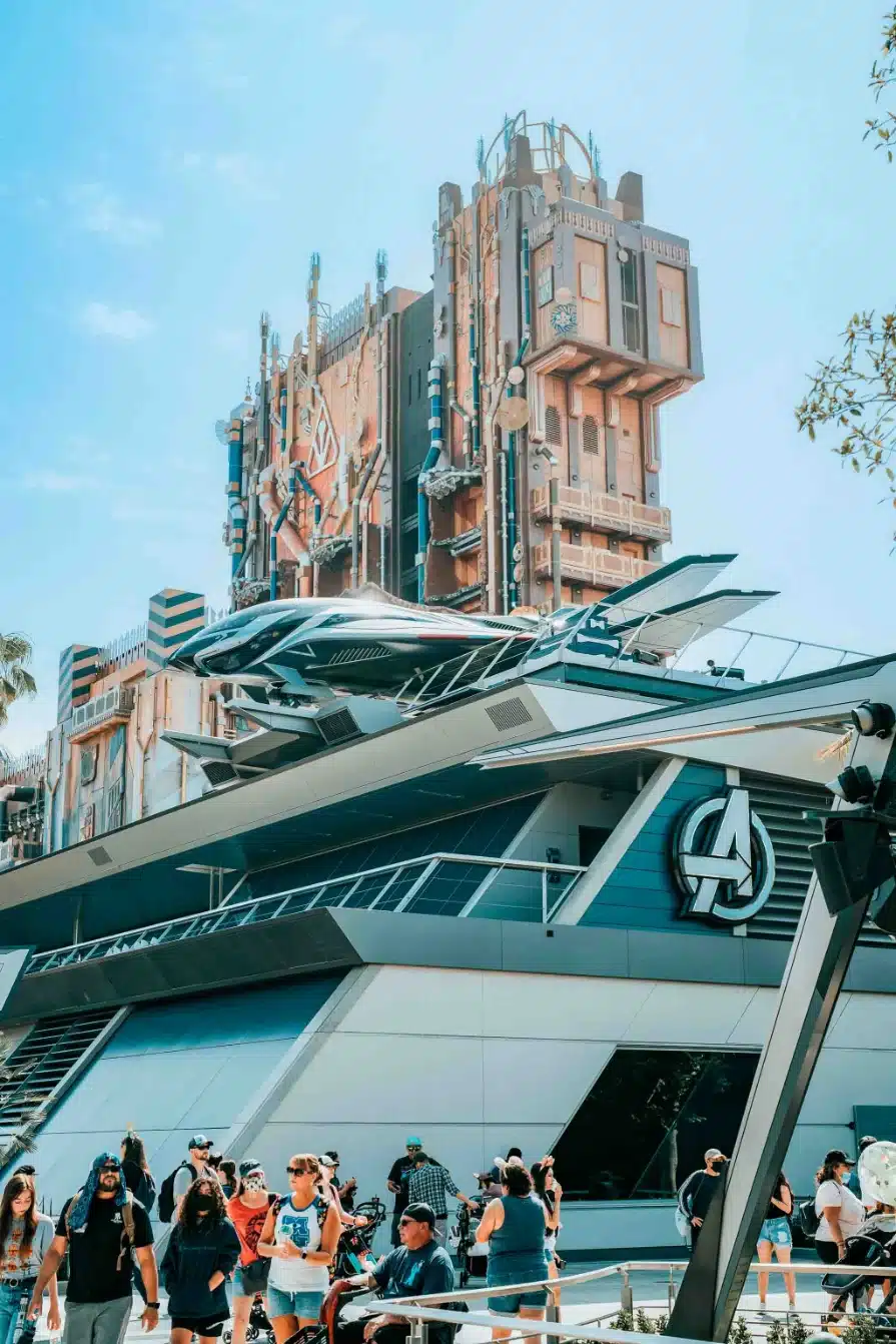 People walking at Avengers Campus with a big Avengers building in the back and a tall abstract building even further featuring Guardians of the Galaxy.
