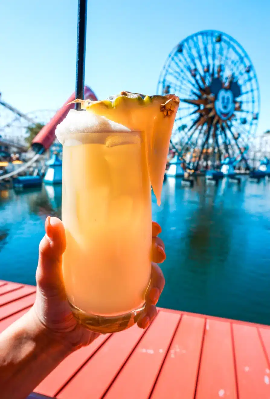 A hand holding a glass of Dole Whip Cocktail at DCA in front of the lagoon and Pixar Pier Ferris wheel.