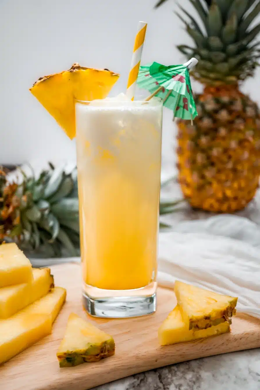 A tall glass of Dole Whip Cocktail with a pineapple wedge on the glass rim, a white and yellow striped straw, and green umbrella. The glass is sitting on a wooden board with pineapple wedges.