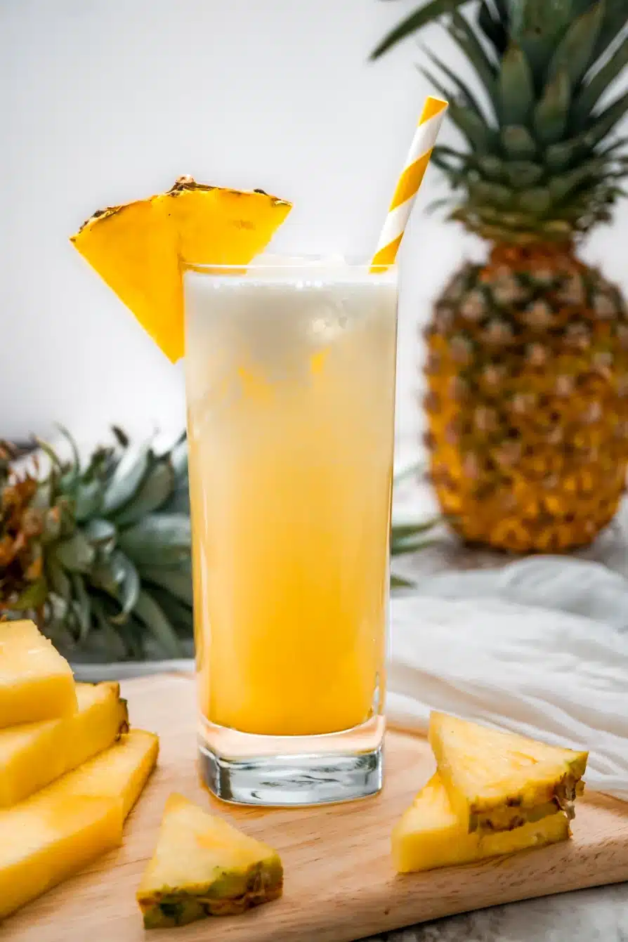 A tall glass of Dole Whip Cocktail with a pineapple wedge on the glass rim and a white and yellow striped straw. The glass is sitting on a wooden board with pineapple wedges.