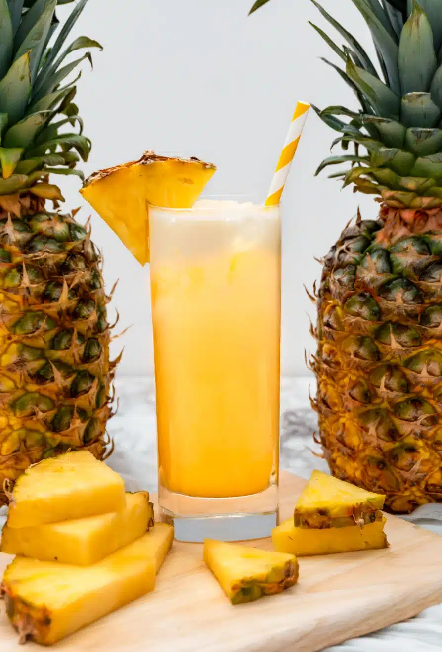 A tall glass of Dole Whip Cocktail with a pineapple wedge on the glass rim between two fresh pineapples.