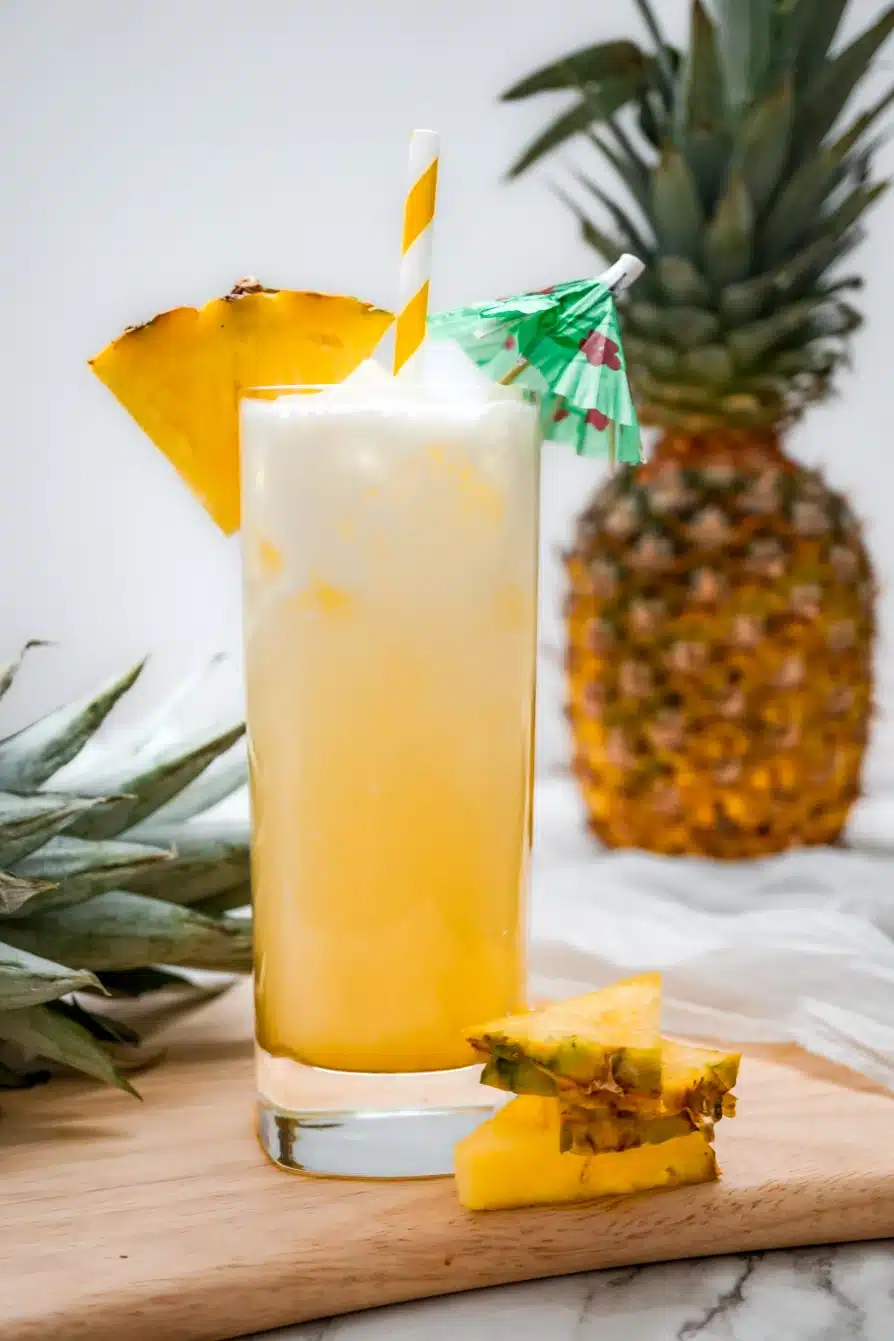 A tall glass of Dole Whip Cocktail with a pineapple wedge on the glass rim, a white and yellow striped straw, and green umbrella. The glass is sitting on a wooden board and a pineapple is in the background.