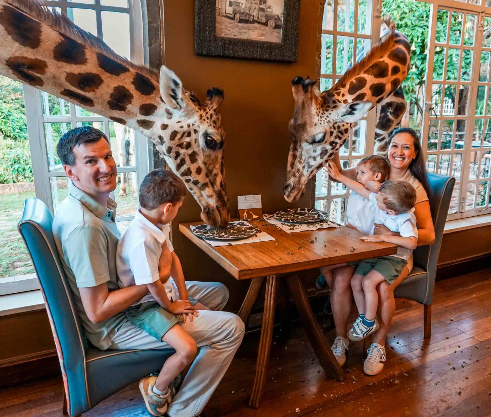 A family of five with three young boys enjoying a classic breakfast with giraffes poking their heads through a window to eat food on a plate—at the Giraffe Manor.