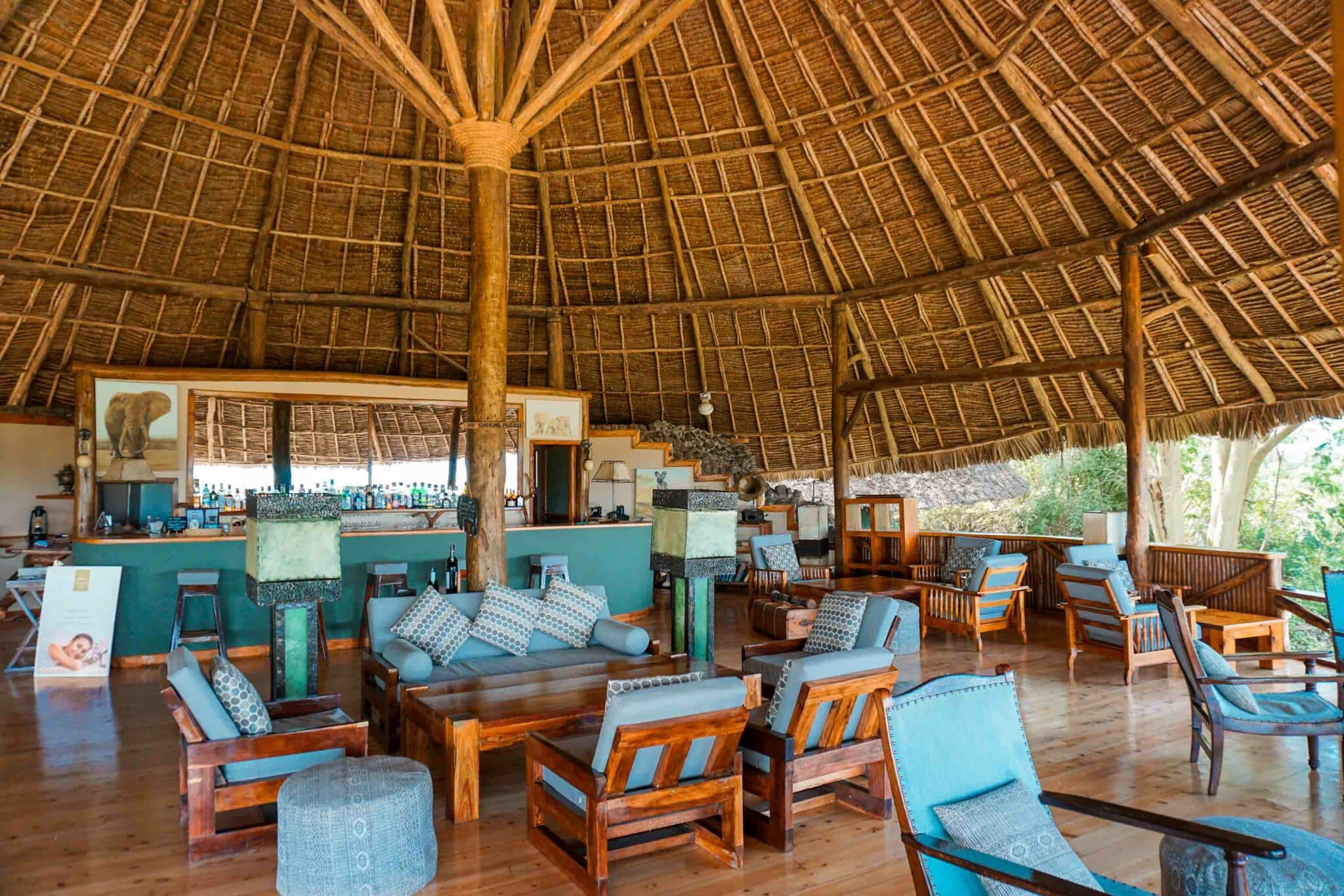 A bar with a commons area for guests to lounge with nice wooden furniture and blue cushions at Elewana Tortilis Camp Amboseli.
